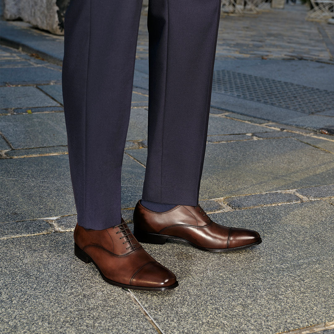 Man in a navy suit pants wearing To Boot's Aidan Brown Cap Toe Oxford standing on a cobblestone city street.