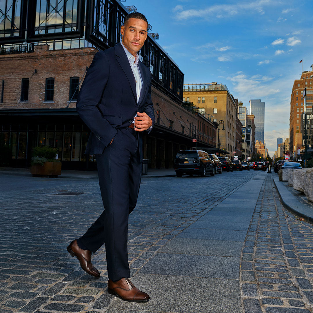 Man in a navy suit wearing To Boot's Aidan Brown Cap Toe Oxford walking on a cobblestone city street with buildings in the background.