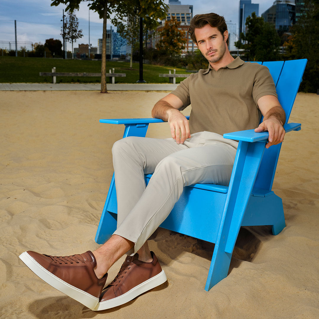 Slide Man sitting on a blue chair wearing To Boot's James Tan Leather Stretch Lace Luxury Low-Top Sneaker in an outdoor setting with a cityscape in the background