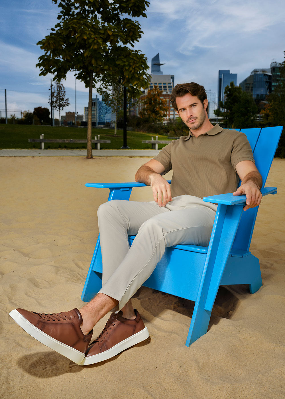 Man sitting on a blue chair wearing To Boot's James Tan Leather Stretch Lace Luxury Low-Top Sneaker in an outdoor setting with a cityscape in the background