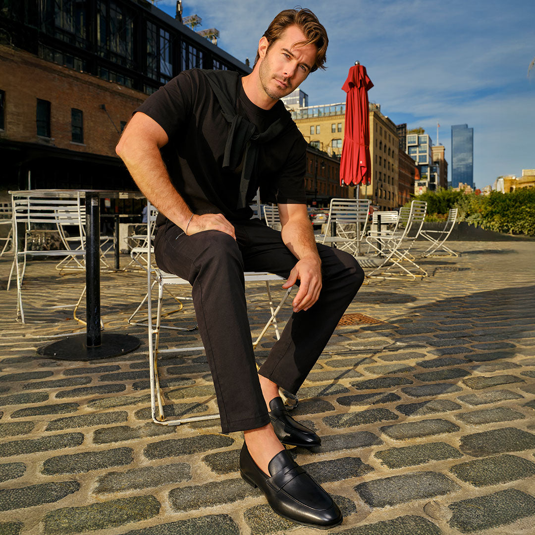 Man in a black outfit wearing To Boot's Keats Black Calf Leather Loafer sitting on a metal chair at an outdoor café on a cobblestone street.