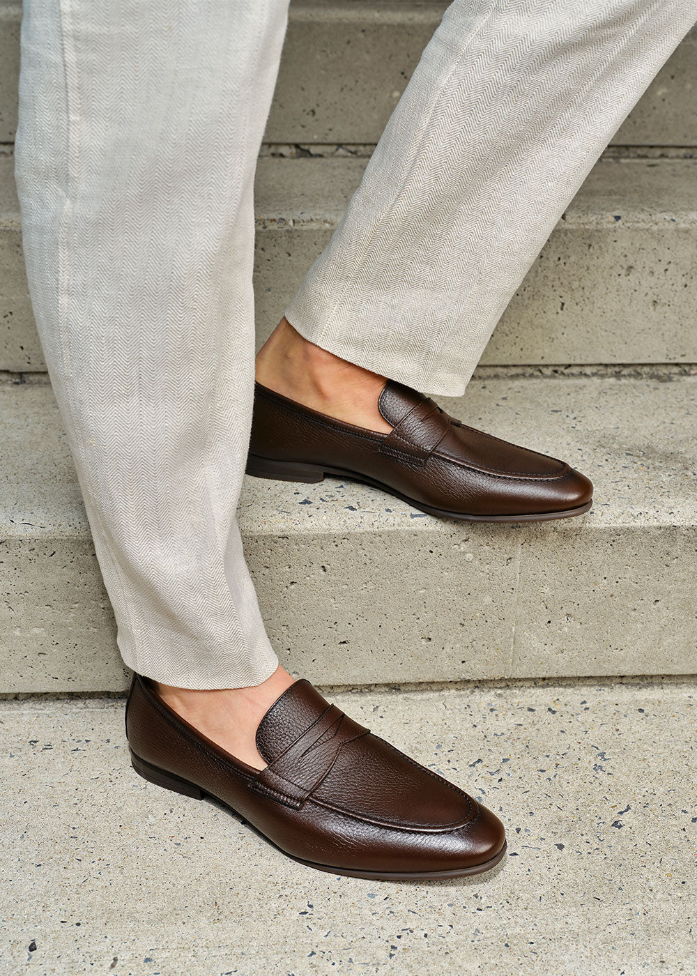 To Boot's Merritt Brown Deer Penny Loafer Slip-On worn with light gray pants on a concrete step.